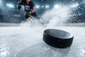 Eishockey-Action: Puck gleitet über das Eis mit aufspritzendem Eisschnee und Spieler im Hintergrund – Hockeyweb.de News